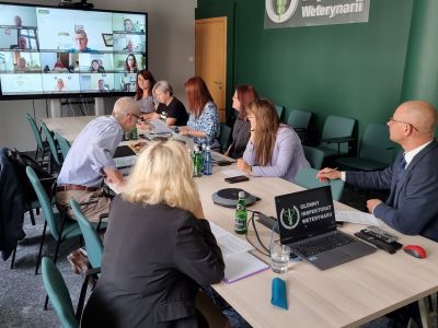 Zdjęcie przedstawia osoby biorące udział w spotkaniu siedzące przy stole patrzące się na ekran na którym są wyświetleni inni uczestnicy spotkania.